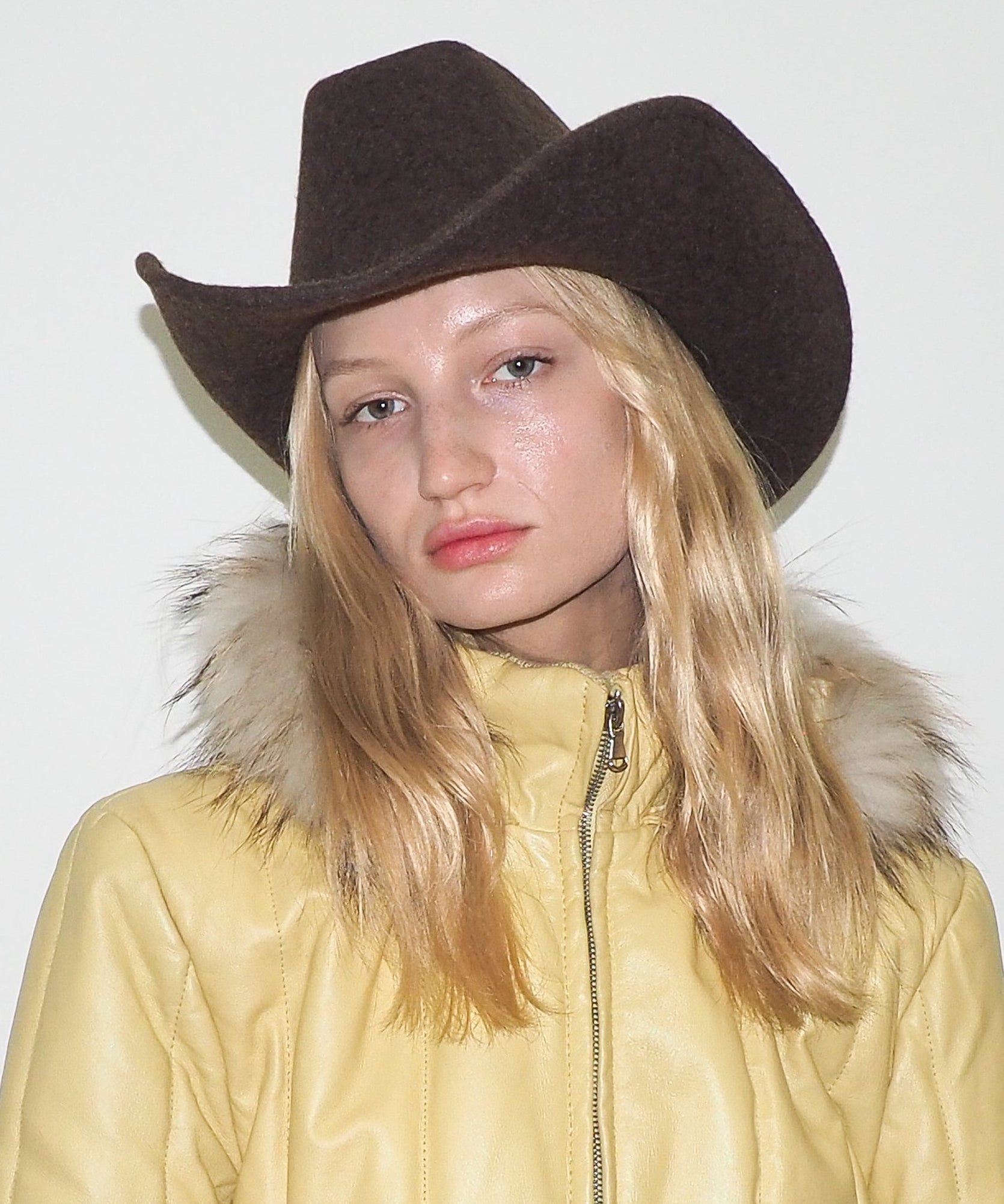 Person wearing a brown cowboy hat and yellow coat with fur hood against a white background