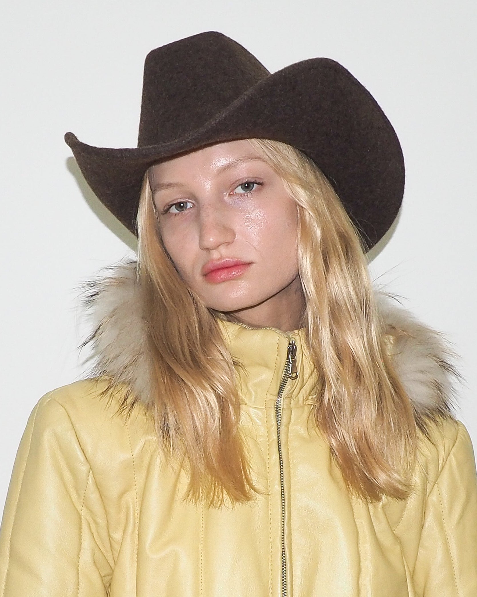 Person wearing a brown cowboy hat and yellow coat with fur hood against a white background
