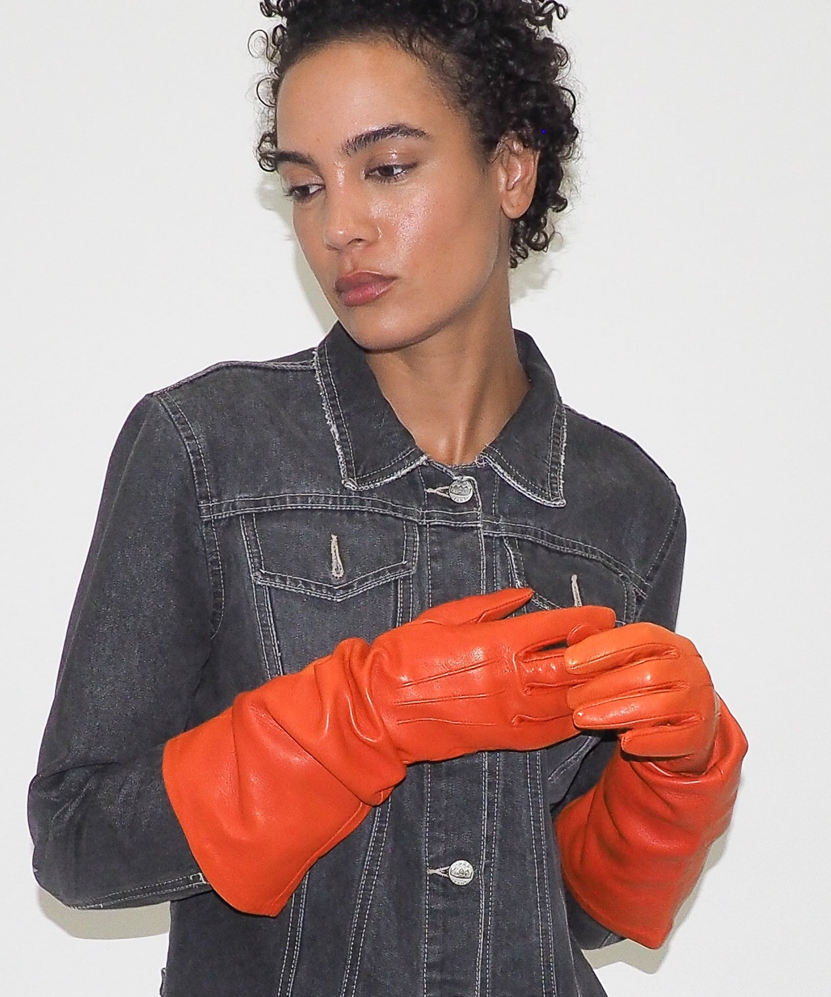 Person wearing a black denim jacket and orange leather gloves against a white background