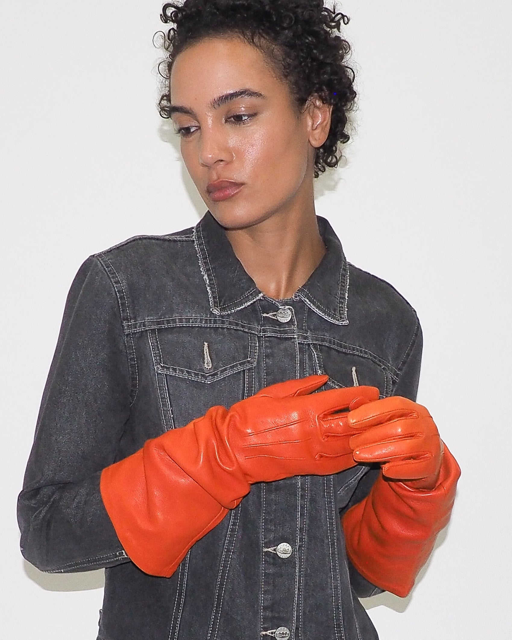 Person wearing a black denim jacket and orange leather gloves against a white background