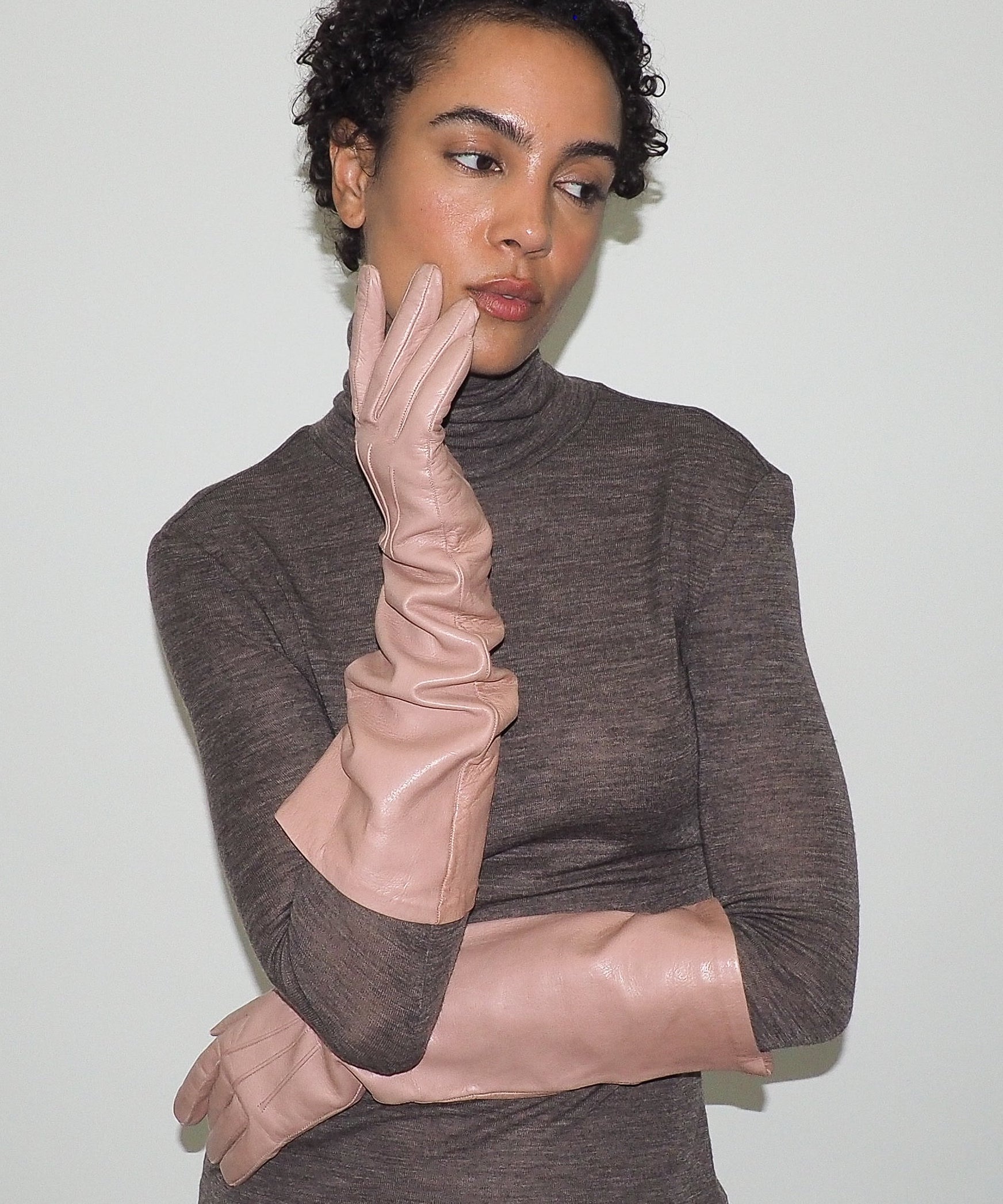 Woman wearing a brown turtleneck sweater and beige gloves against a plain background