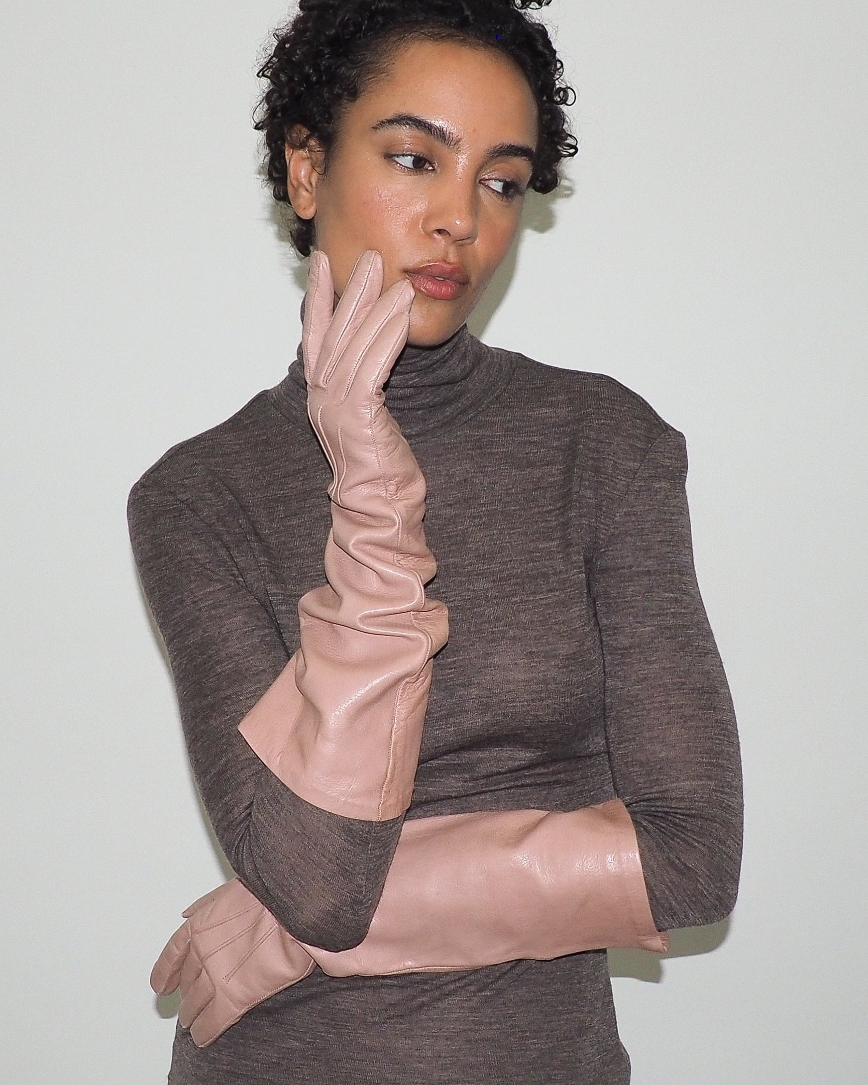 Woman wearing a brown turtleneck sweater and beige gloves against a plain background