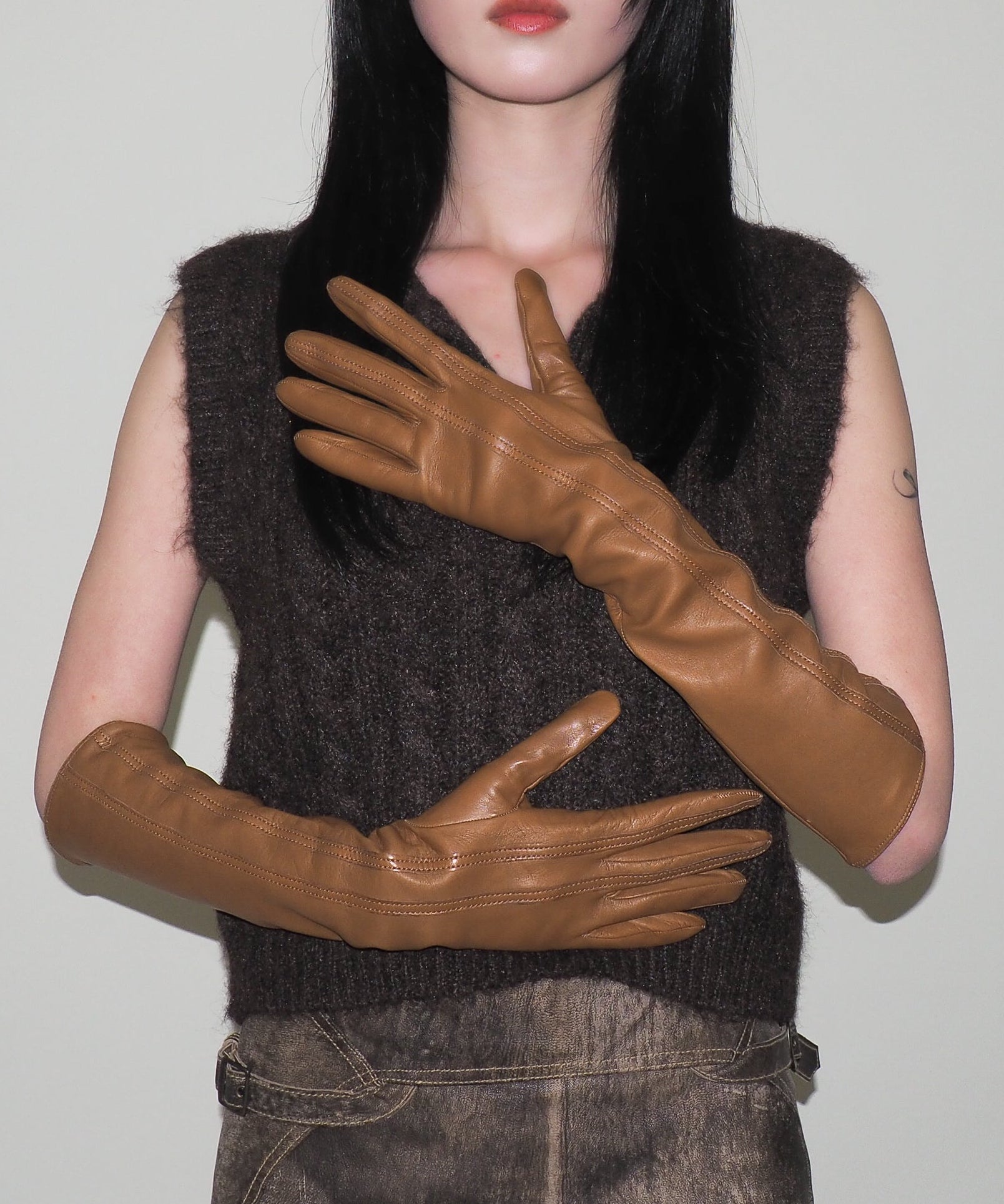 Person wearing a dark brown sleeveless top and brown leather gloves against a plain background