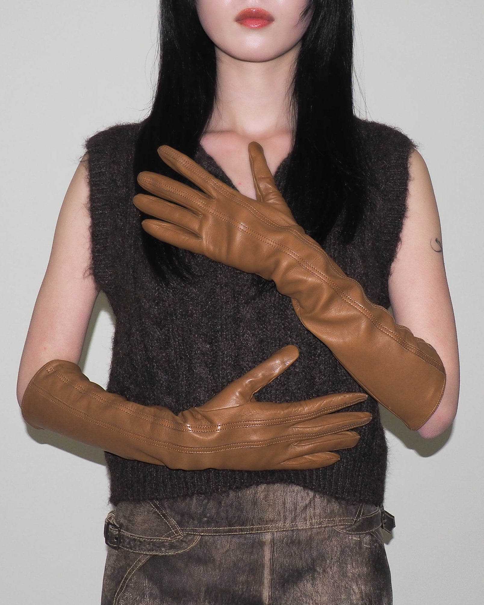 Person wearing a dark brown sleeveless top and brown leather gloves against a plain background