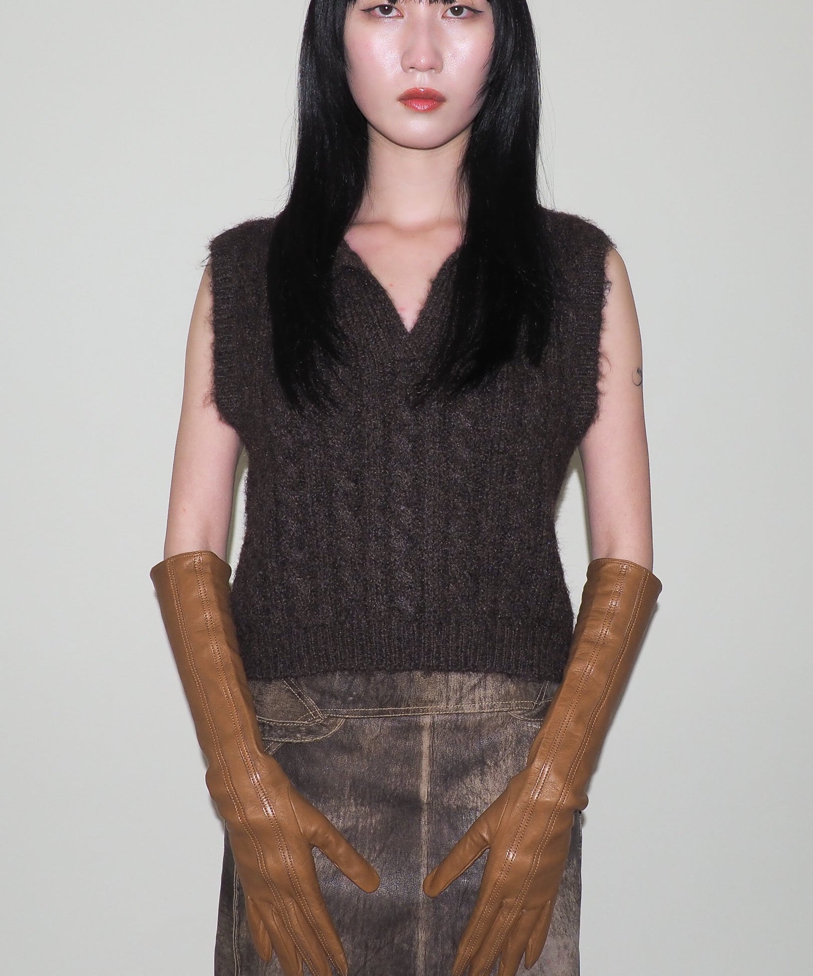 Person wearing a dark brown sleeveless knitted top and brown leather gloves against a plain background