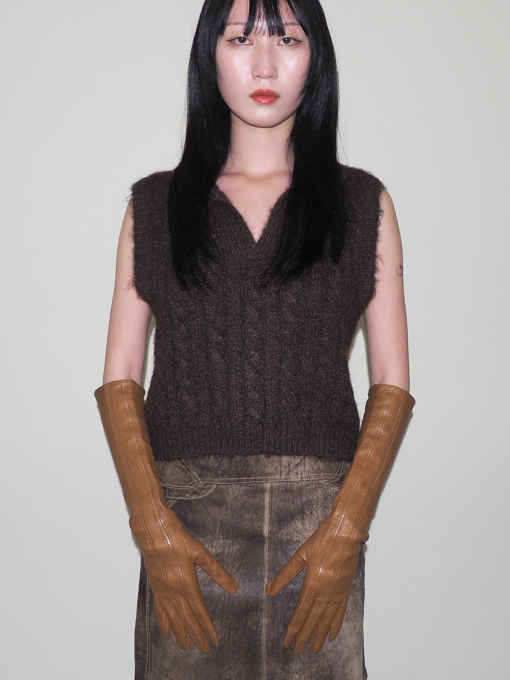 Person wearing a dark brown sleeveless knitted top and brown leather gloves against a plain background