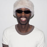 Person wearing a white beanie with fur trim and dark sunglasses, smiling against a plain background.