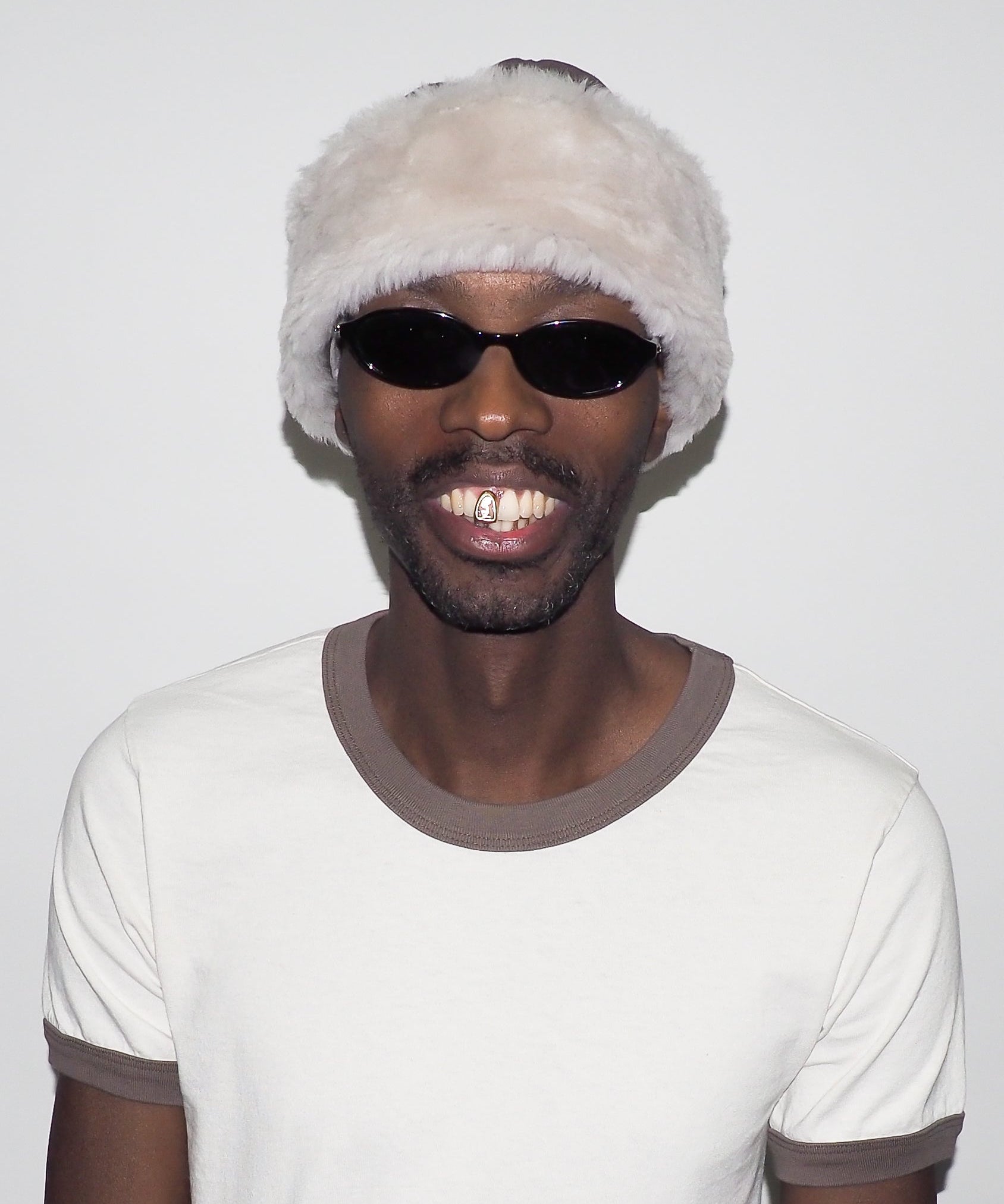 Person wearing a white beanie with fur trim and dark sunglasses, smiling against a plain background.