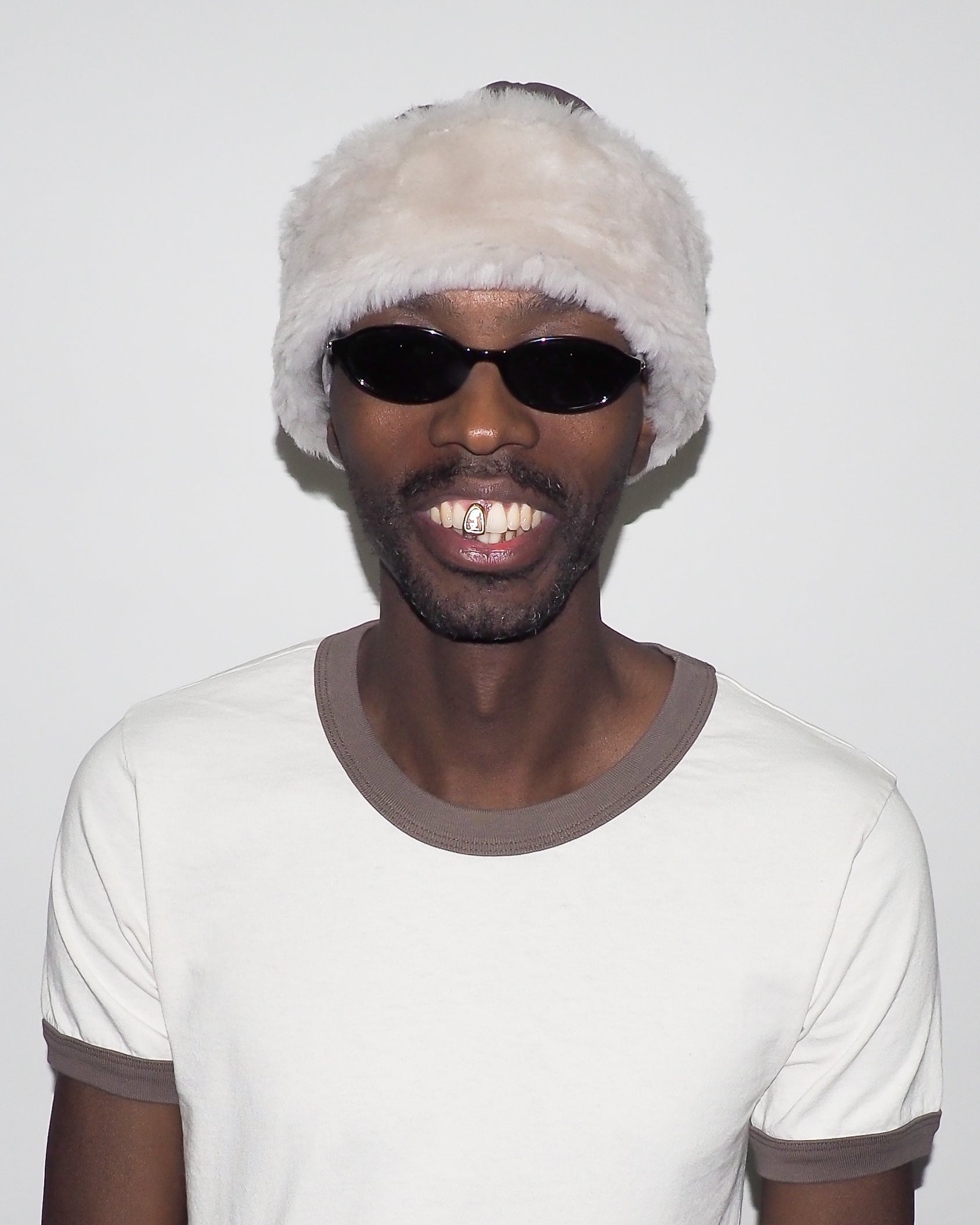 Person wearing a white beanie with fur trim and dark sunglasses, smiling against a plain background.