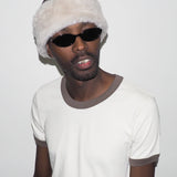 Person wearing a white beanie with fur trim and sunglasses on a plain background
