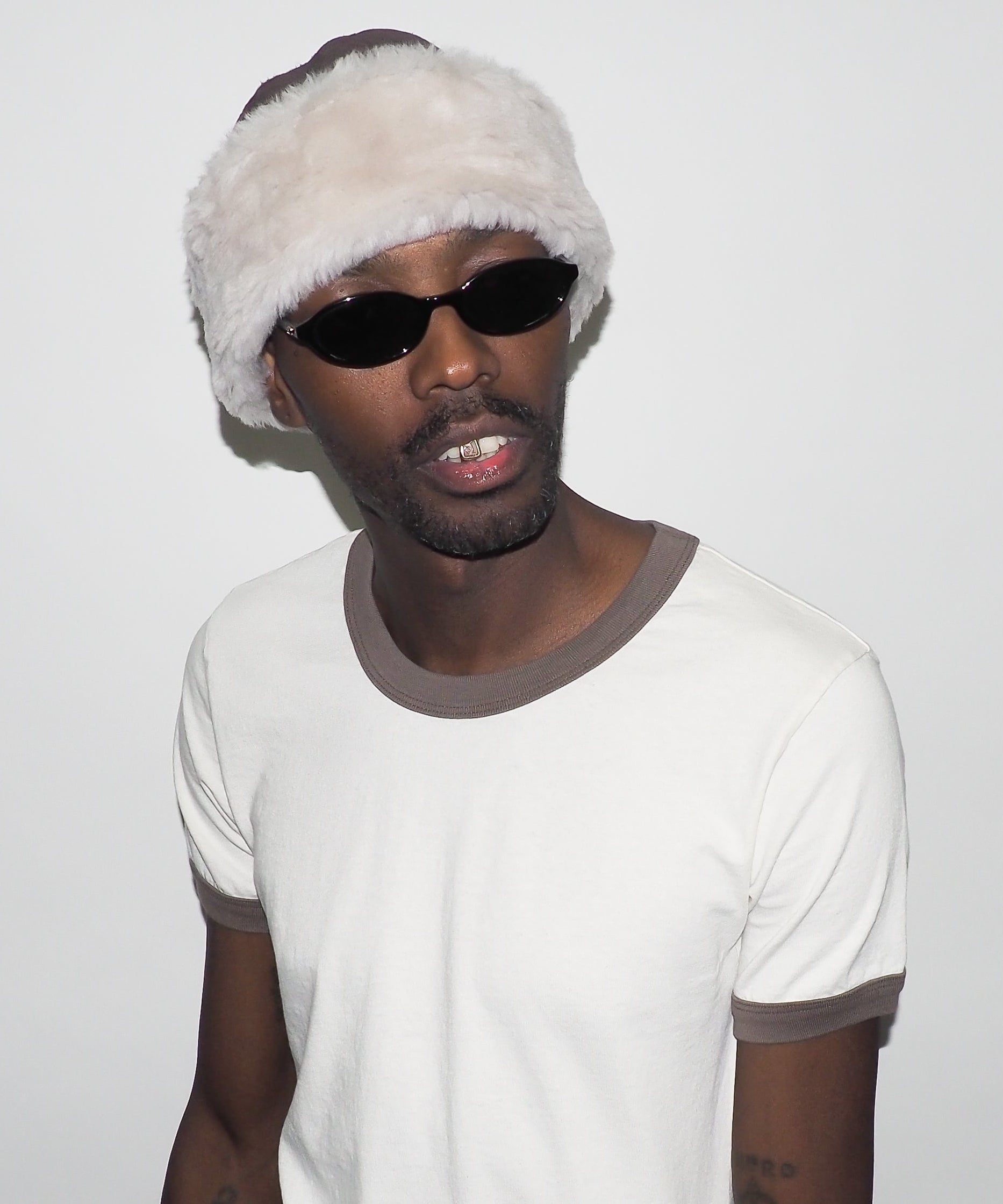Person wearing a white beanie with fur trim and sunglasses on a plain background