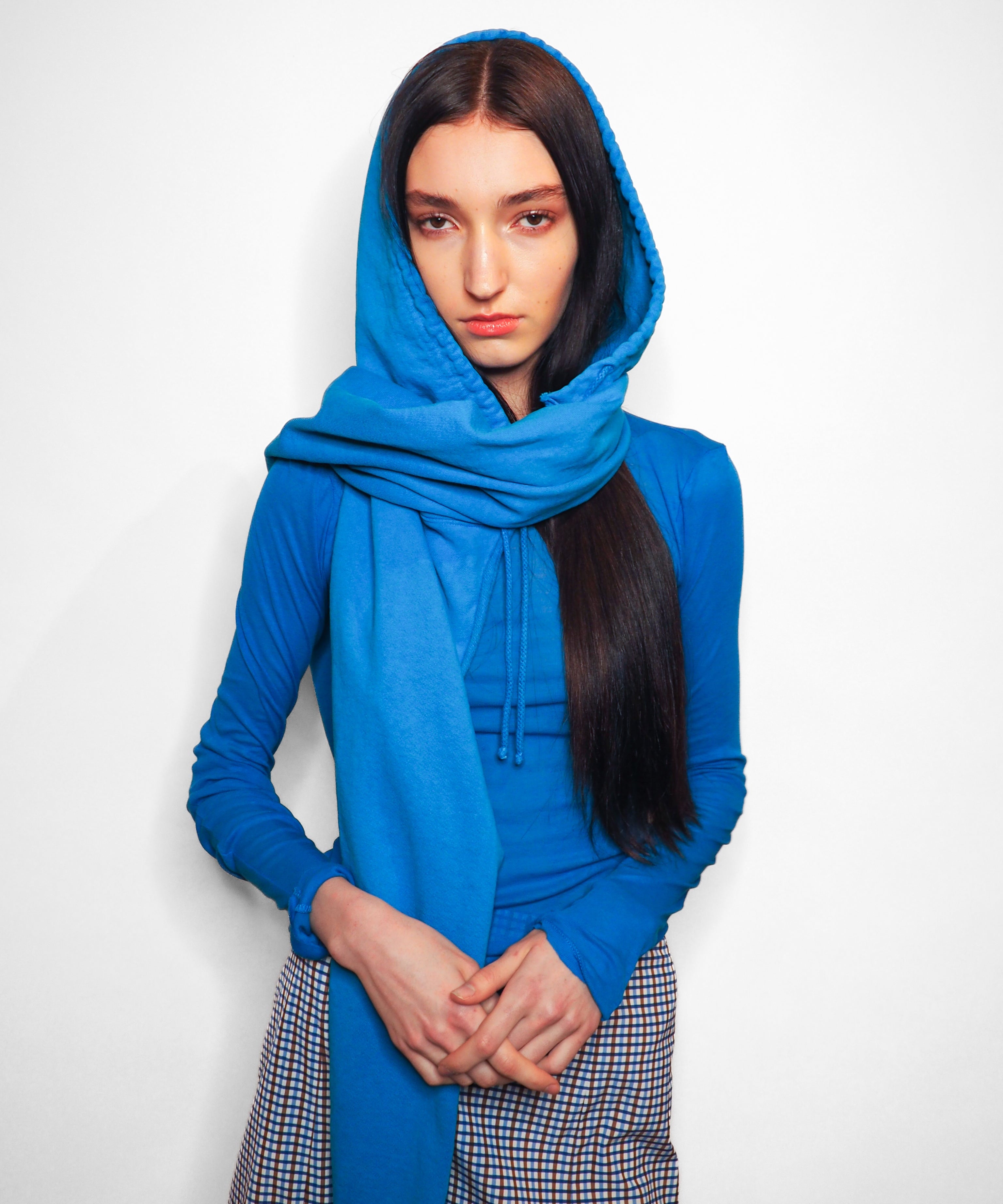 A model wearing a bright blue hooded sweatshirt and matching scarf, paired with a blue, white, and black checkered skirt, standing against a plain white background.