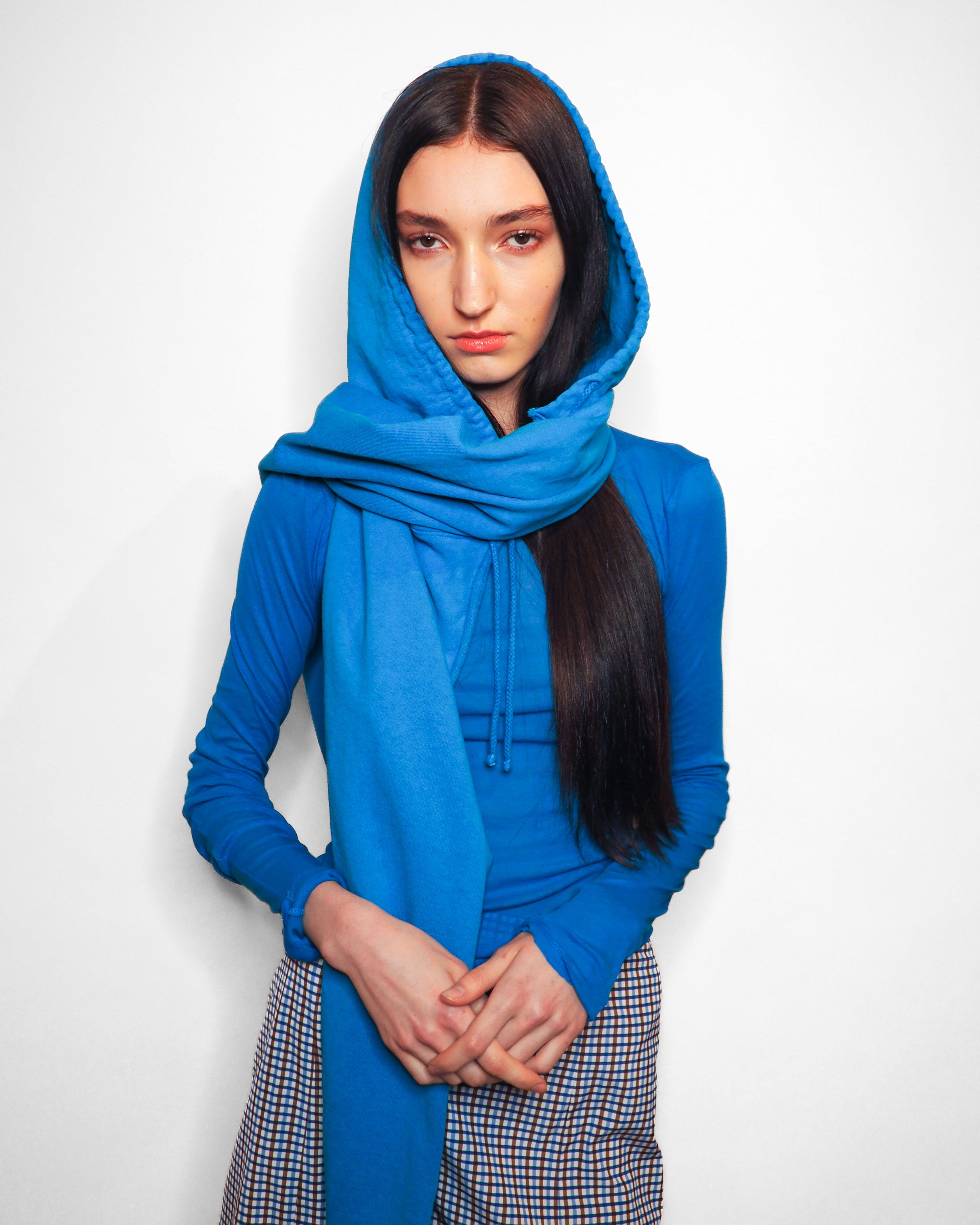 A model wearing a bright blue hooded sweatshirt and matching scarf, paired with a blue, white, and black checkered skirt, standing against a plain white background.