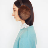 A side-profile view of a model wearing brown fur earmuffs with a dark headband, a light blue knit sweater, and a striped shirt collar, set against a plain white background.