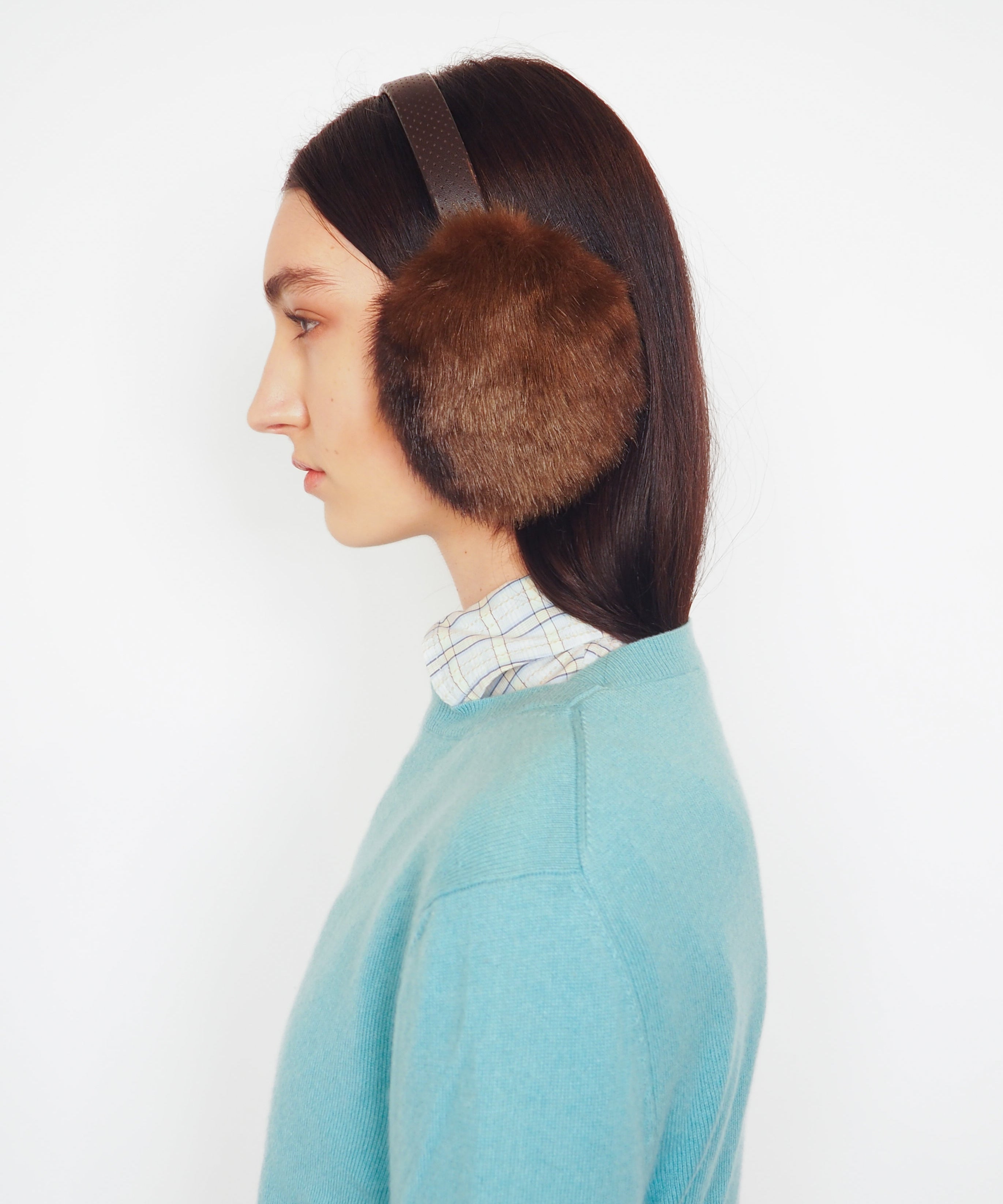 A side-profile view of a model wearing brown fur earmuffs with a dark headband, a light blue knit sweater, and a striped shirt collar, set against a plain white background.