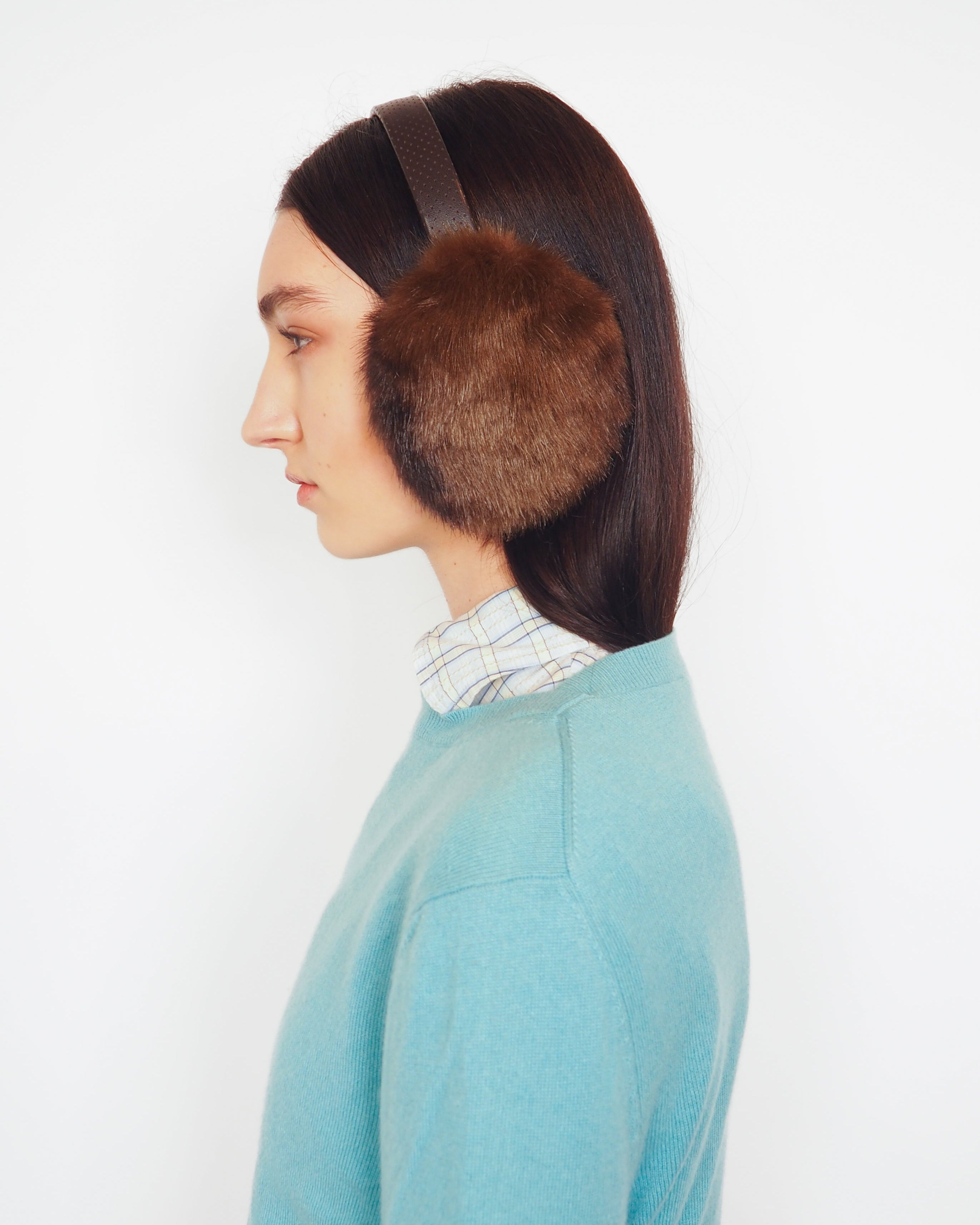 A side-profile view of a model wearing brown fur earmuffs with a dark headband, a light blue knit sweater, and a striped shirt collar, set against a plain white background.