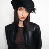 A model wearing a black leather jacket, a dark brown ribbed top, and a black fuzzy beret-style hat with feather trim, posed against a plain white studio background.