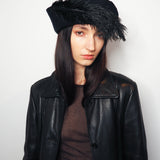 A model wearing a black leather jacket, a dark brown top, and a black feather-trimmed beret hat, photographed against a plain light gray background.