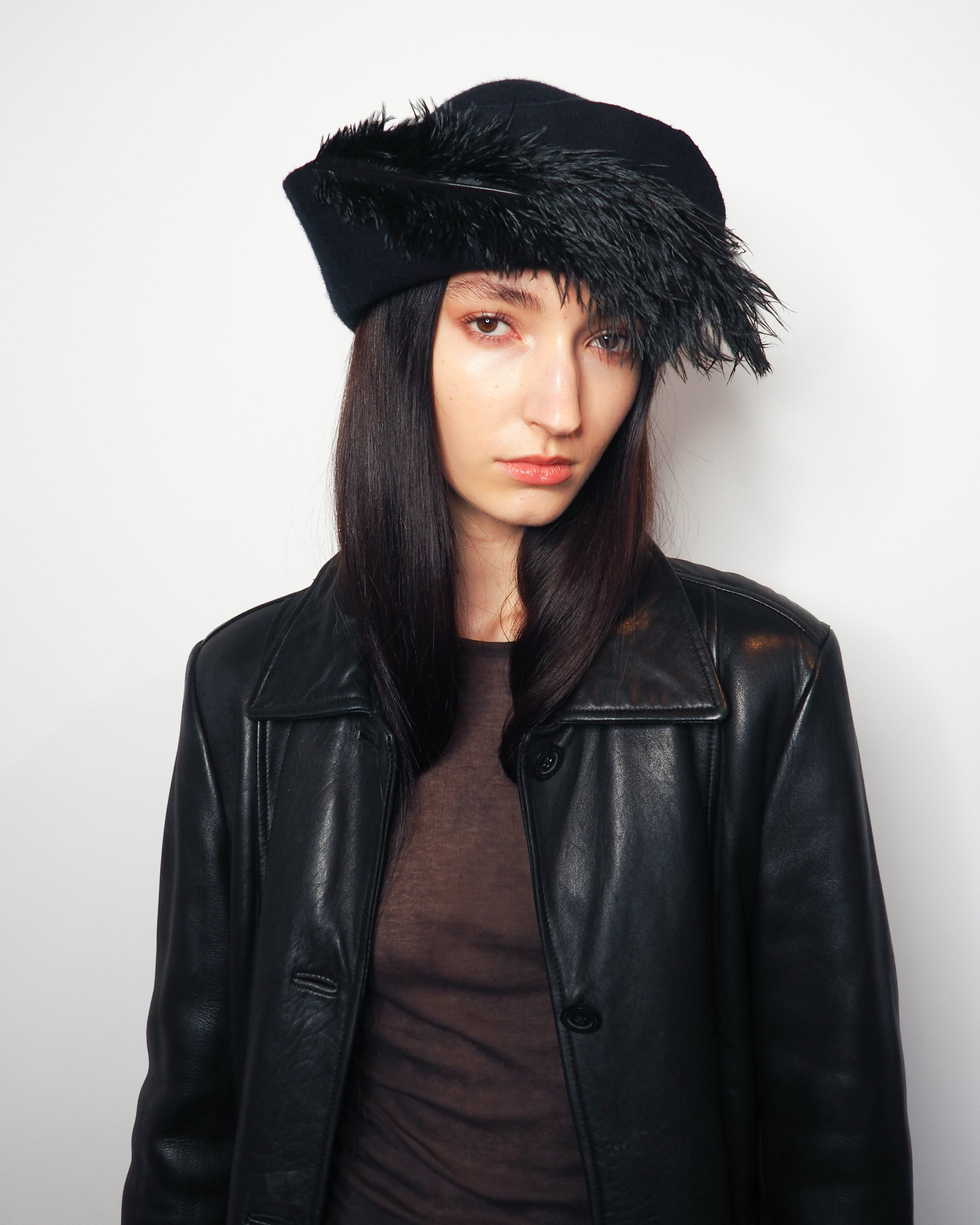 A model wearing a black leather jacket, a dark brown top, and a black feather-trimmed beret hat, photographed against a plain light gray background.