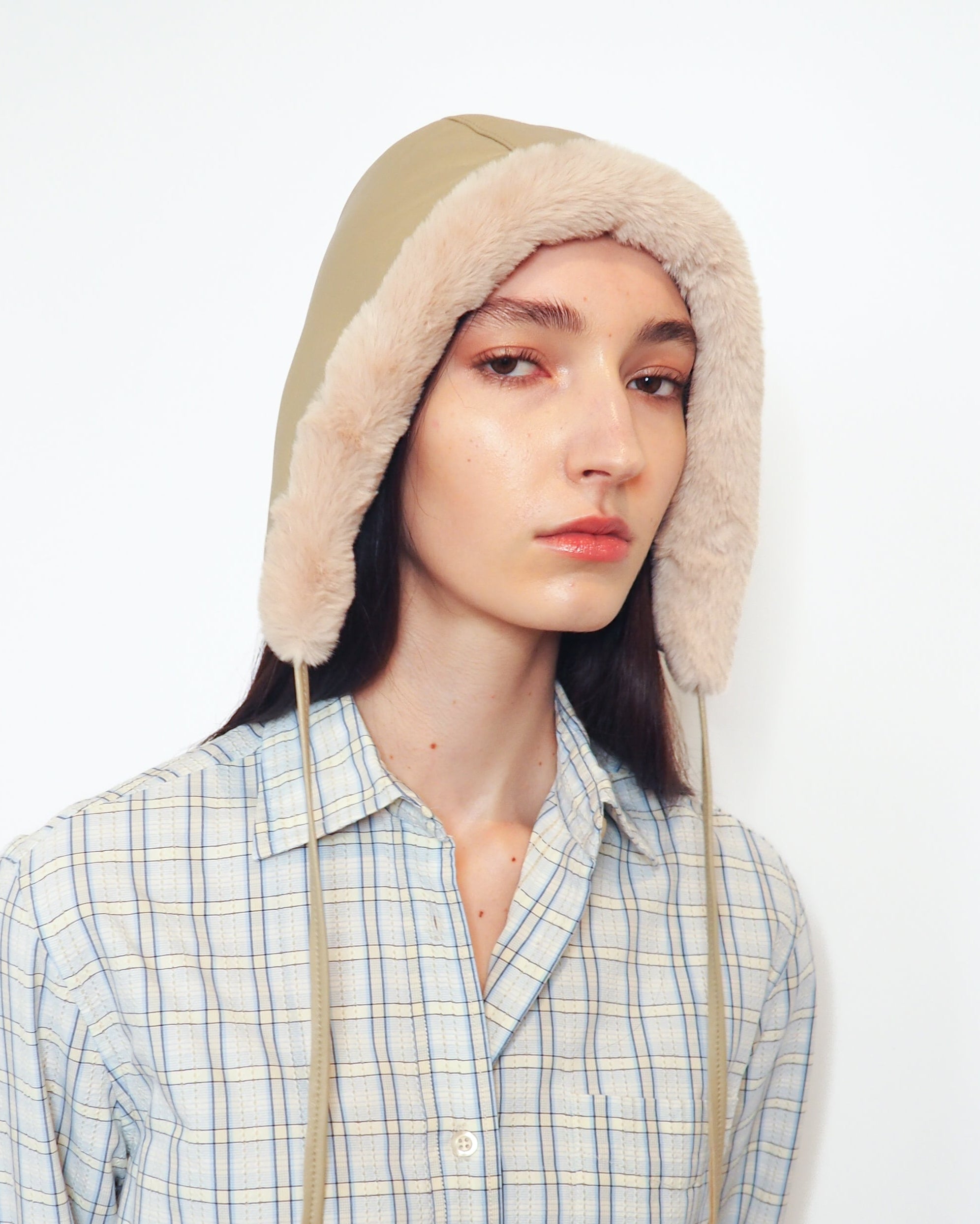 A model wearing a beige and cream plaid button-up shirt with a matching beige knit hood featuring faux-fur trim, set against a plain white background.