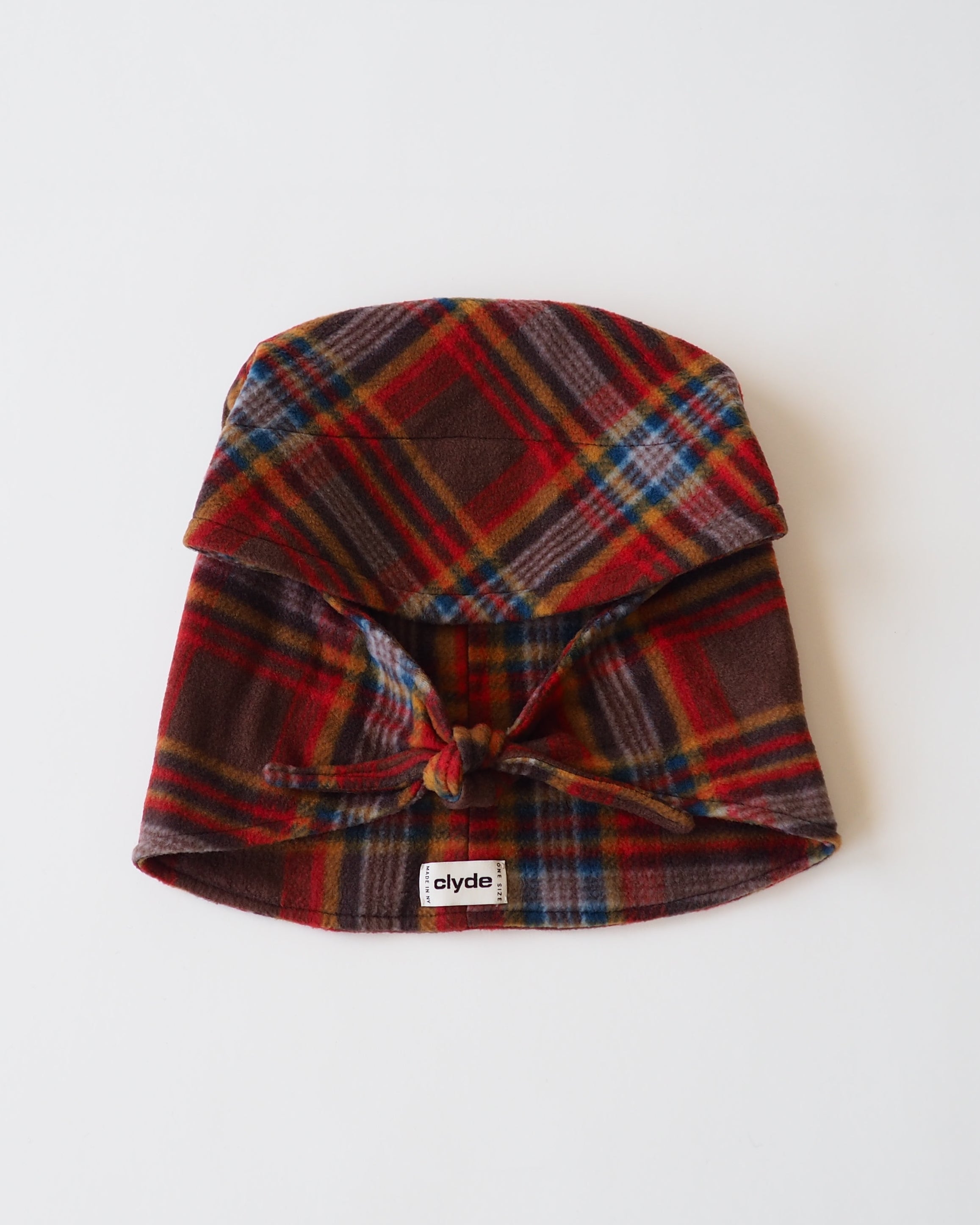 A plaid fabric hat with a tie closure at the front, featuring a red, brown, and blue tartan pattern, laid flat on a white background. A small white tag with the brand name 'clyde' is visible near the bottom.