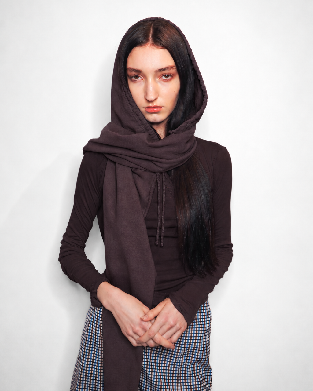 A model wearing a dark brown hooded long-sleeve top and a matching brown scarf, paired with a blue and white checkered skirt, against a plain light gray background.