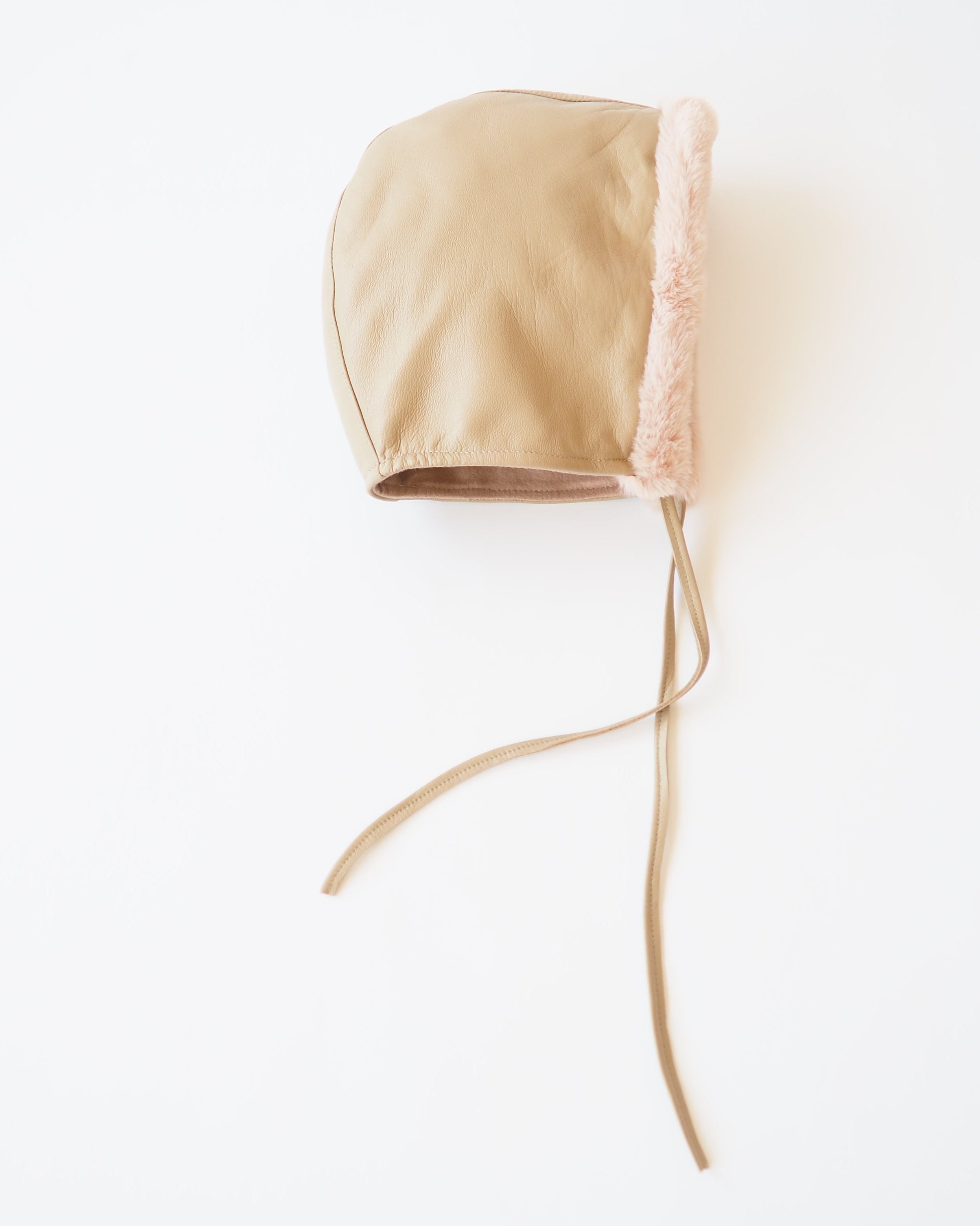 A beige leather bonnet with a light pink faux-fur trim and long beige drawstrings, photographed on a clean white background.